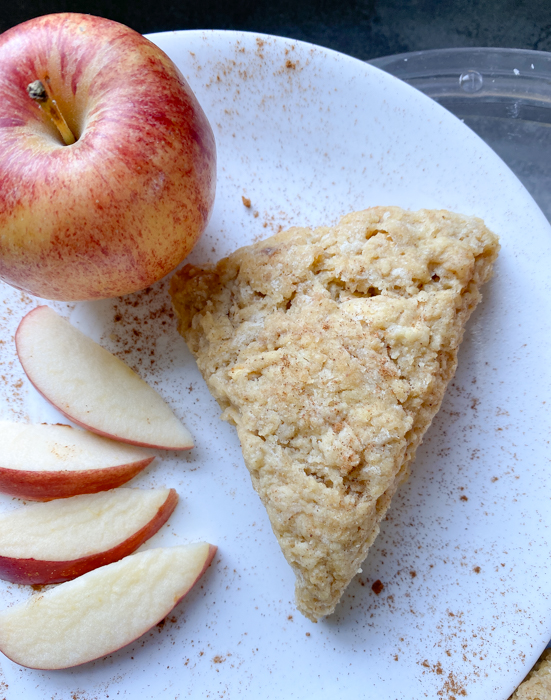 One sourdough einkorn apple scone on a white plate. The plate is sprinkled with cinnamon and includes a sliced and whole apple.