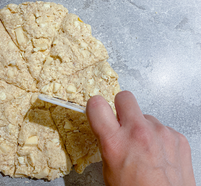 A woman's hand cutting sourdough einkorn scone dough into triangles using a bench scraper.
